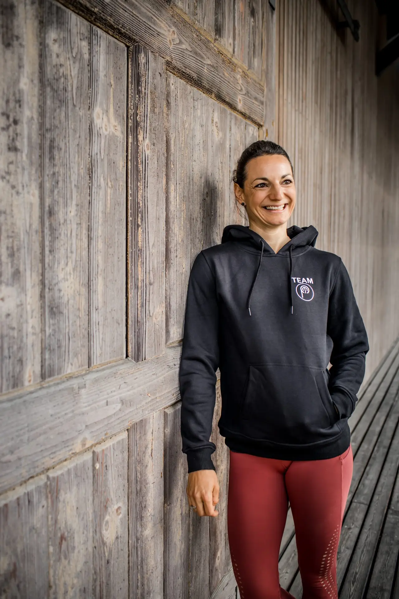 Stefanie Bauer, Heilpraktikerin und Sportwissenschaftlerin, lehnt lächelnd an einer verwitterten Holzwand